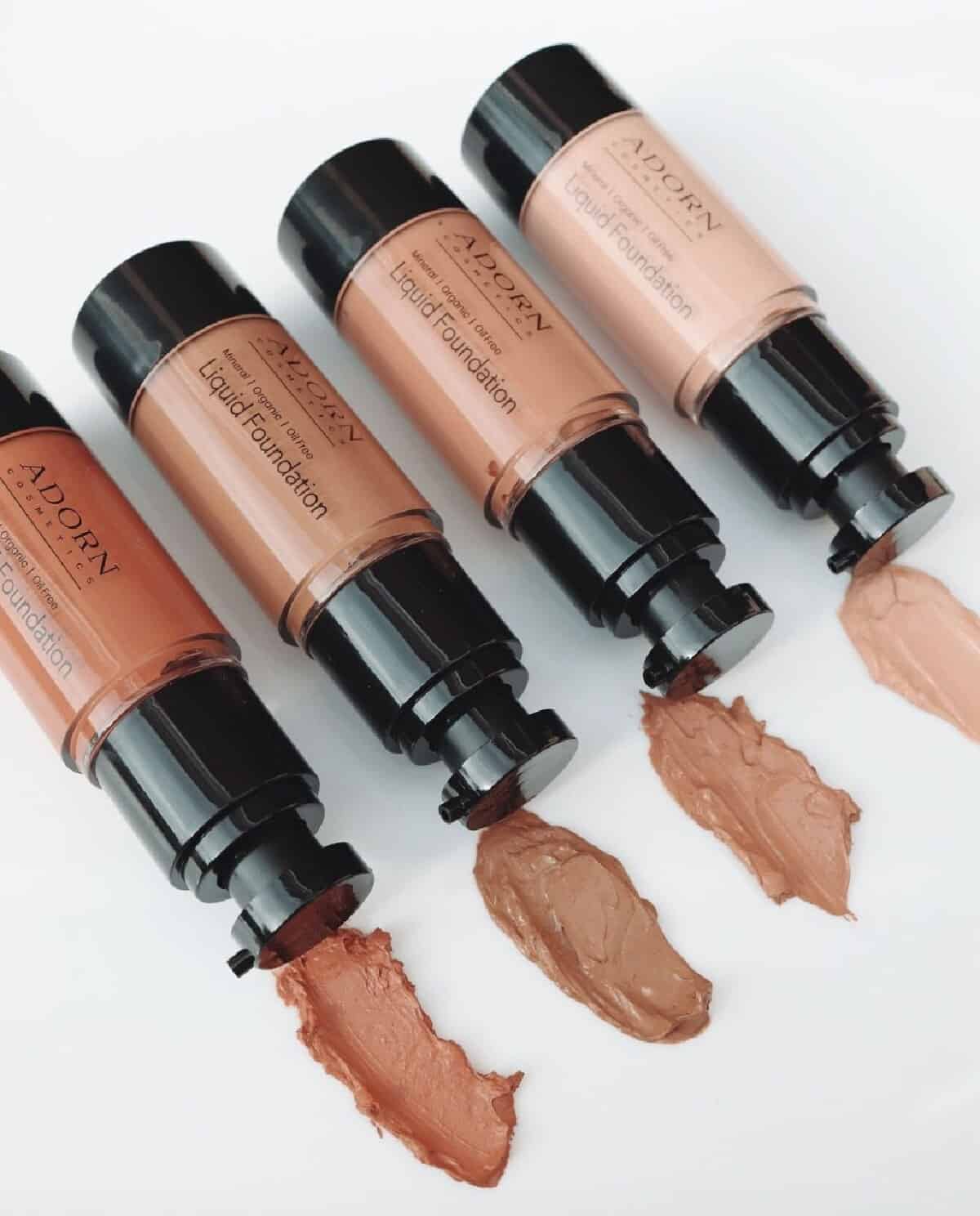 Four bottles of Adorn Cosmetics liquid foundation on a white background laying on their side diagonally with a sample of the color shade shown in front of the dispensing nozzle.