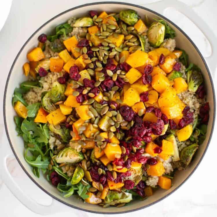 A large serving bowl fill with arugula quinoa salad topped with roasted butternut squash, dried cranberries, and pepitas.