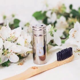 A small recyclable aluminum container containing vegan floss on a white background next to spring flowers and a bamboo toothbrush with black bristles.
