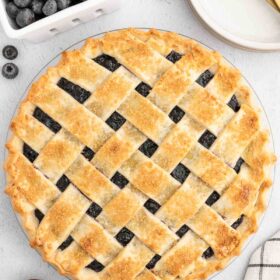 A whole vegan blueberry pie with a golden lattice crust, sitting next to a carton of blueberries.