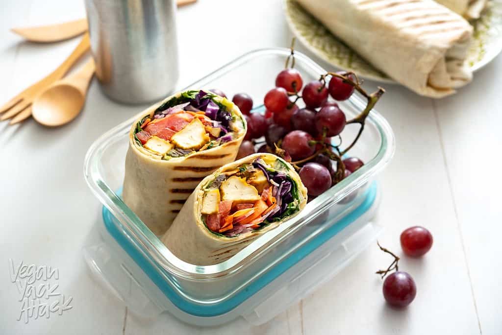 grilled chipotle tofu wraps in a glass container with a bunch of red grapes. the wraps have tofu and veggies like cabbage, greens, carrots, and tomatoes