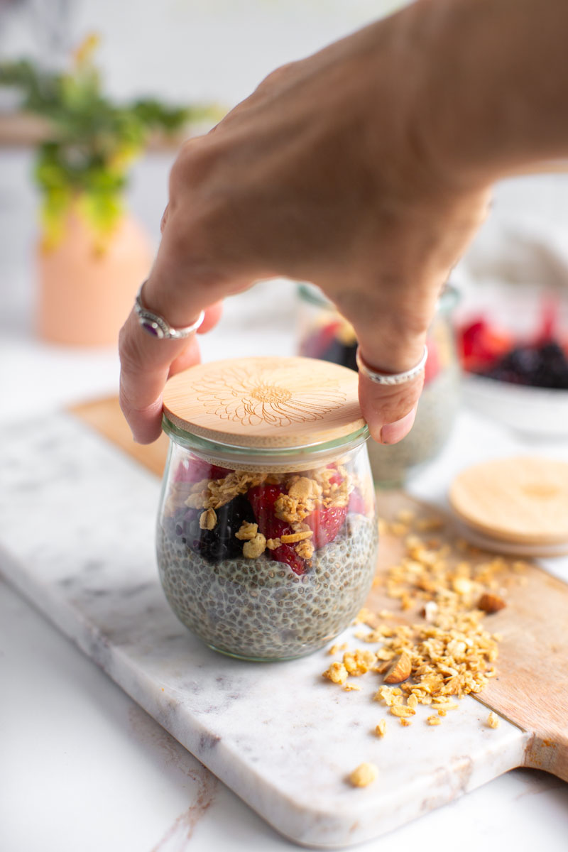 A hand placing a bamboo lid on a small glass jar filled with chia pudding.