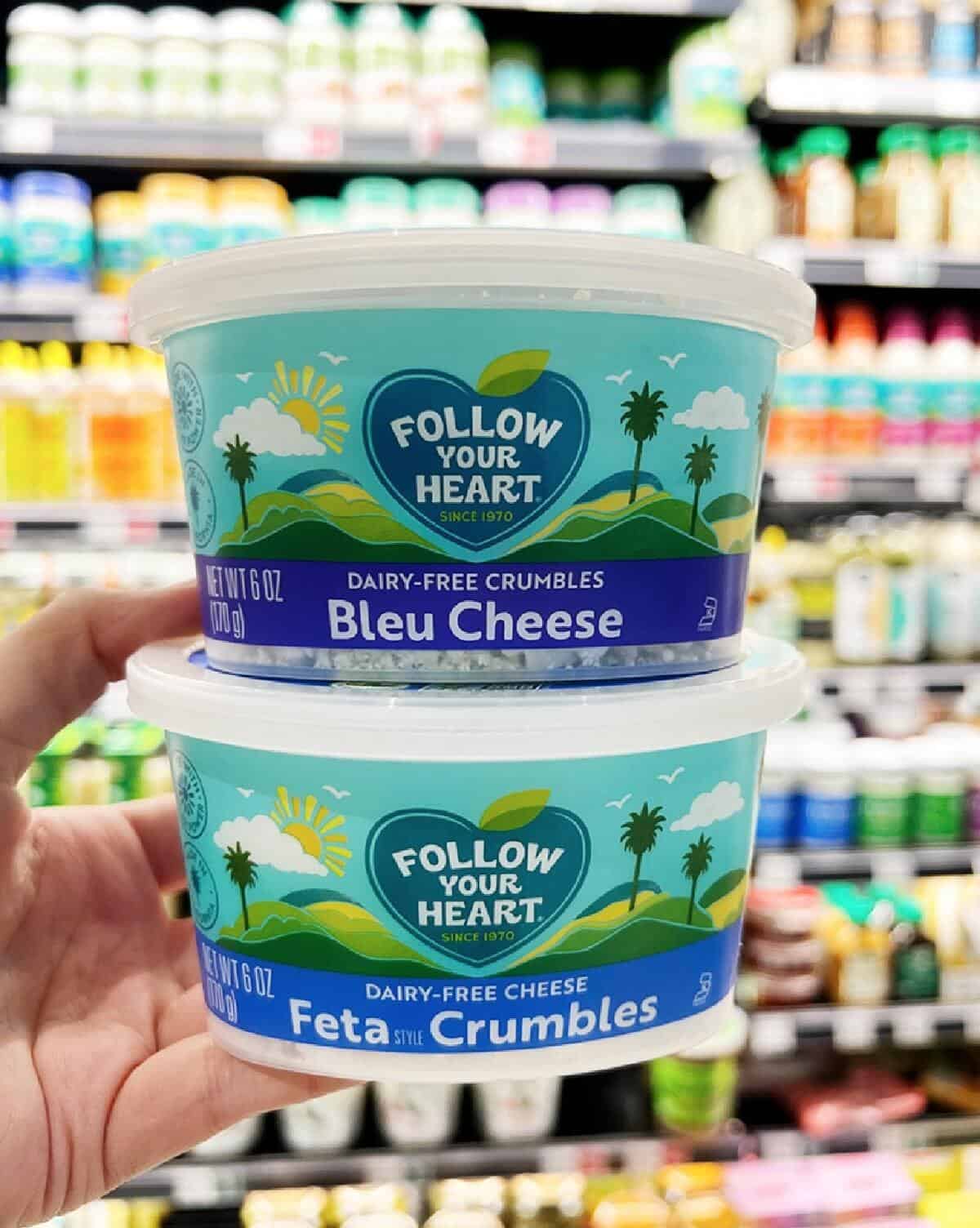 A hand holding two plastic tubs of Follow Your Heart Vegan Blue Cheese crumbles against a grocery store shelf background.