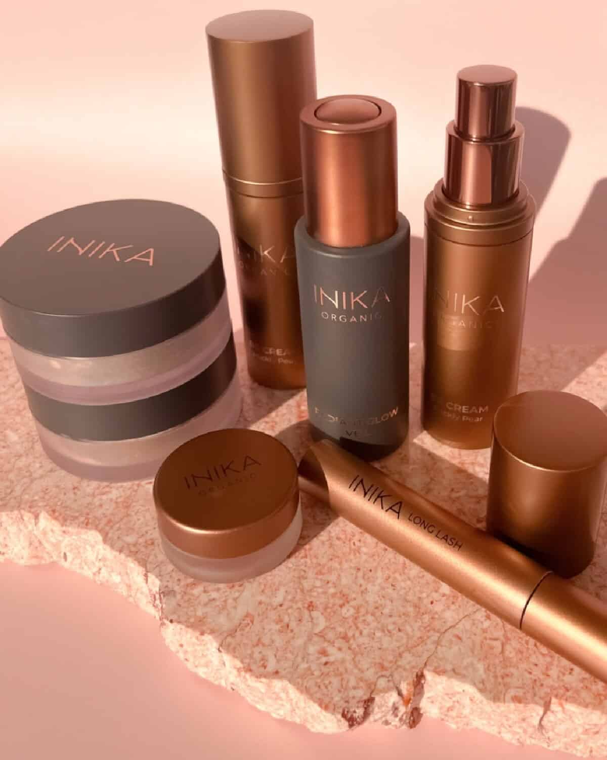 A collection of copper colored bottles and containers from the Inika Organic line of make up products displayed on a rose pink background on top of a pink-colored stone slab.