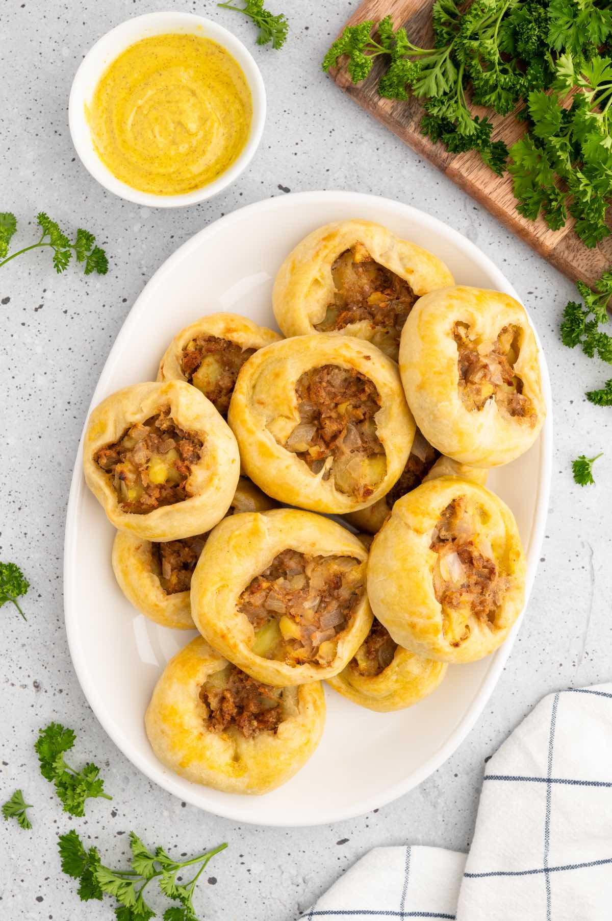 A white serving platter piled with vegan potato knish pastries garnished with parsley and served with a side of mustard.