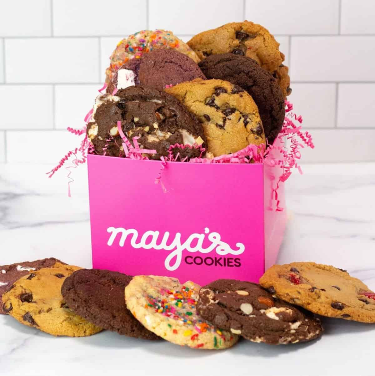 Pink box with the Maya's Cookies logo on it, with cookies sticking up out of it and laying around it.