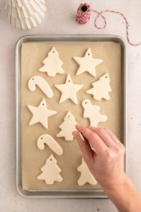 Holiday shaped salt cough ornaments on baking sheet with a hand hovering over to add the holes with a wooden stick.