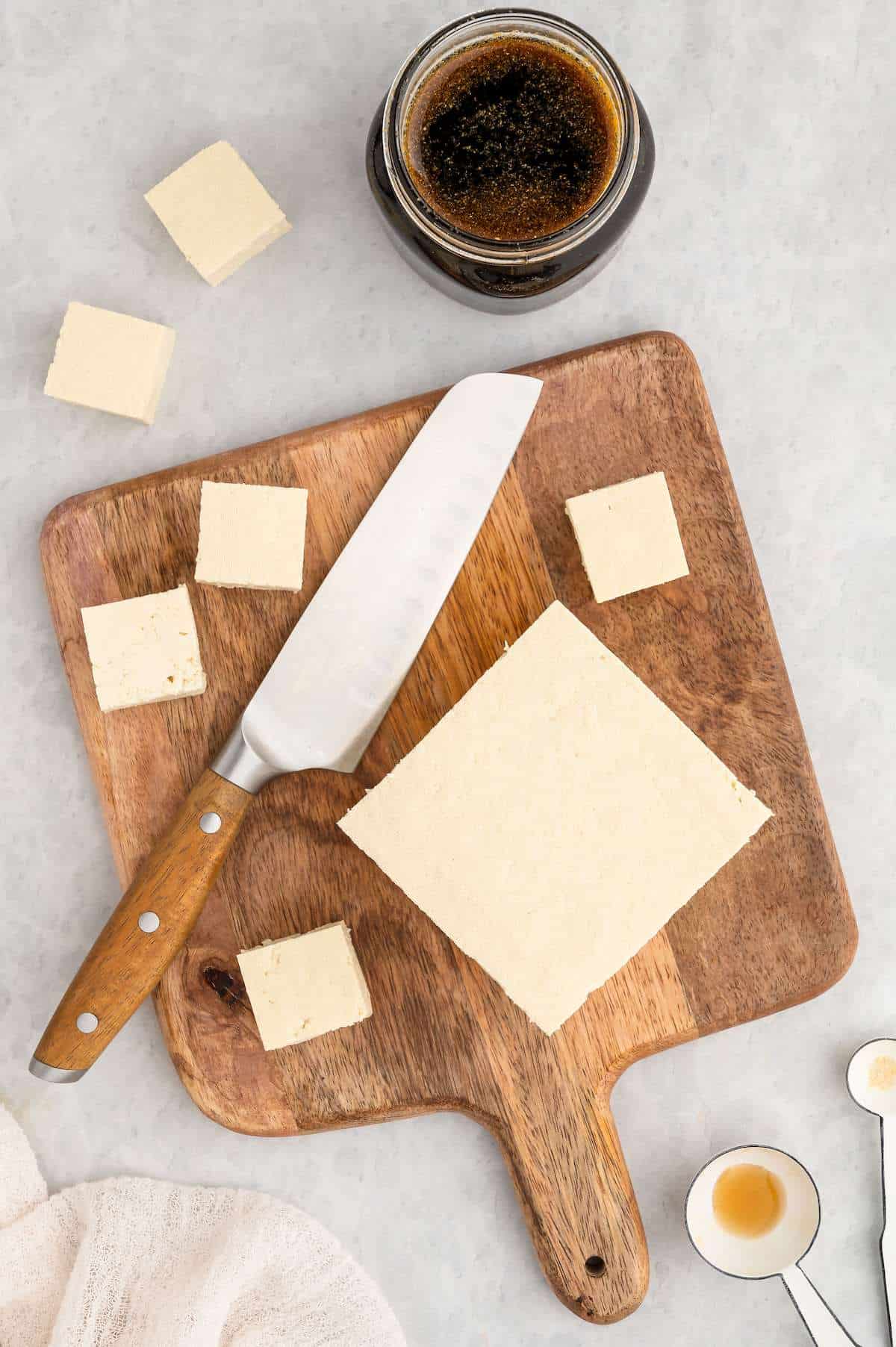 Tofu on a cutting board with a knife, partially sliced into cubes.
