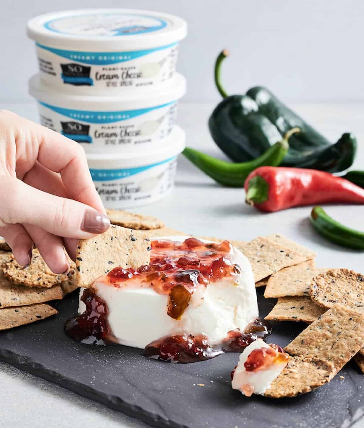 A stack of So Delicious plant-based cream cheese behind of platter of vegan cream cheese spread with crackers.