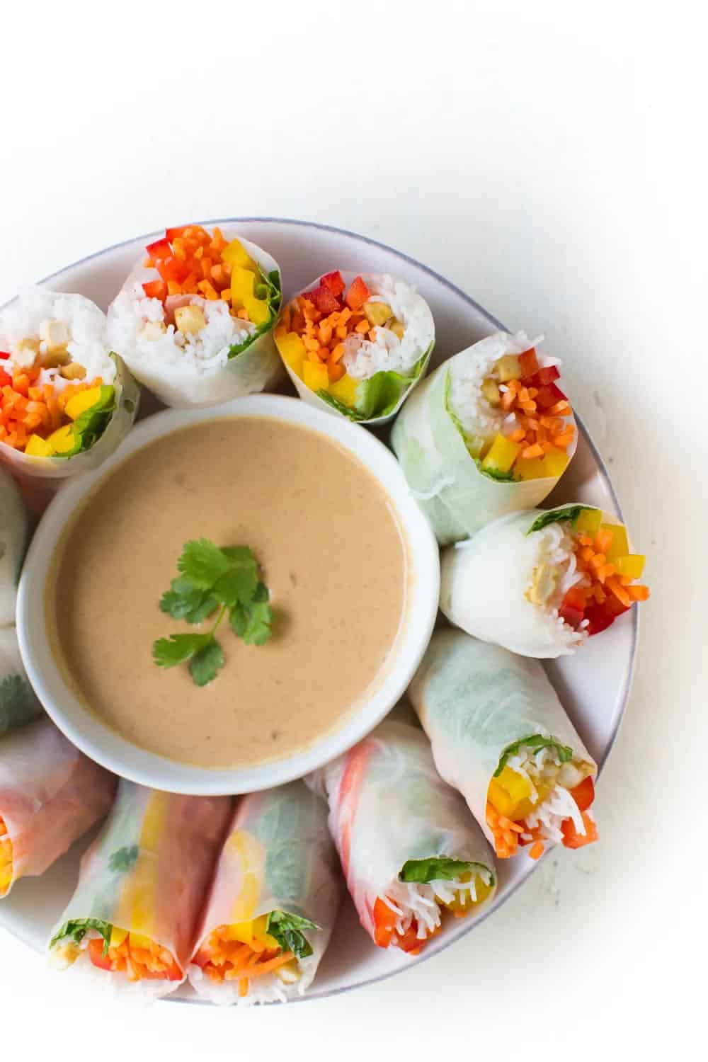fresh spring rolls filled with tofu, lettuce, red and yellow bell pepper strips, vermicelli noodles, and shredded carrots on a plate with a bowl of peanut sauce in the middle with cilantro garnish