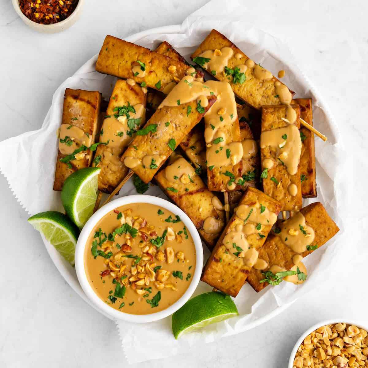 Tofu satay skewers on a serving platter with peanut dipping sauce and lime wedges.