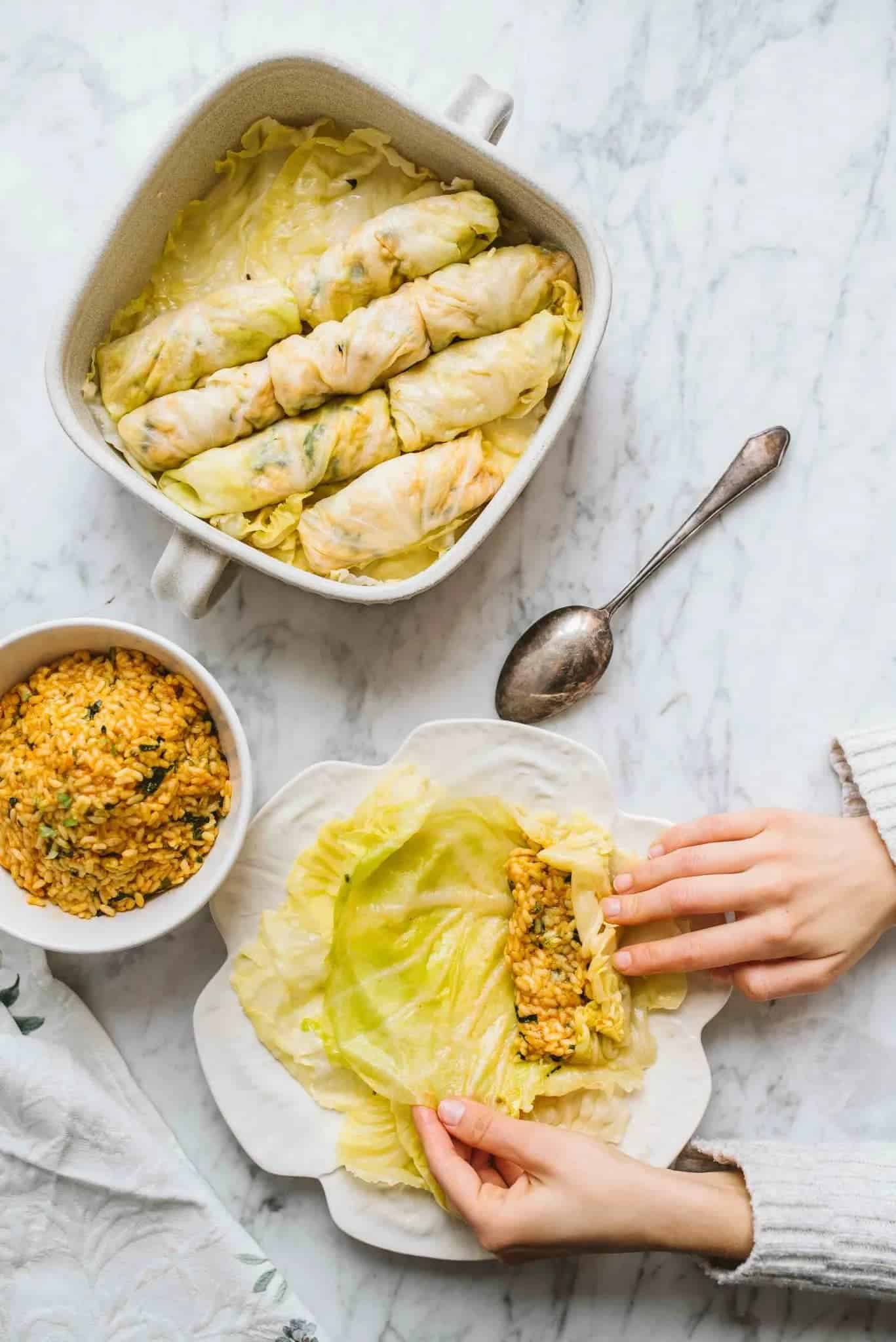 vegan cabbage rolls in a dish with light-skinned hands in the bottom right corner assembling one filled with rice and spices