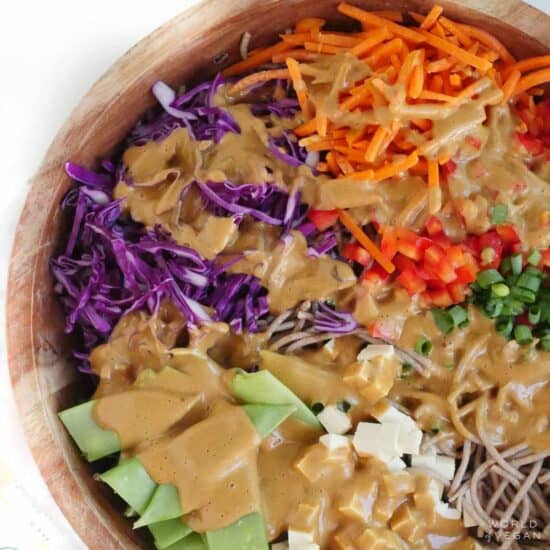 A bowl of cold peanut noodle salad with purple cabbage, carrots, peas, tofu, green onion, and sesame peanut sauce.