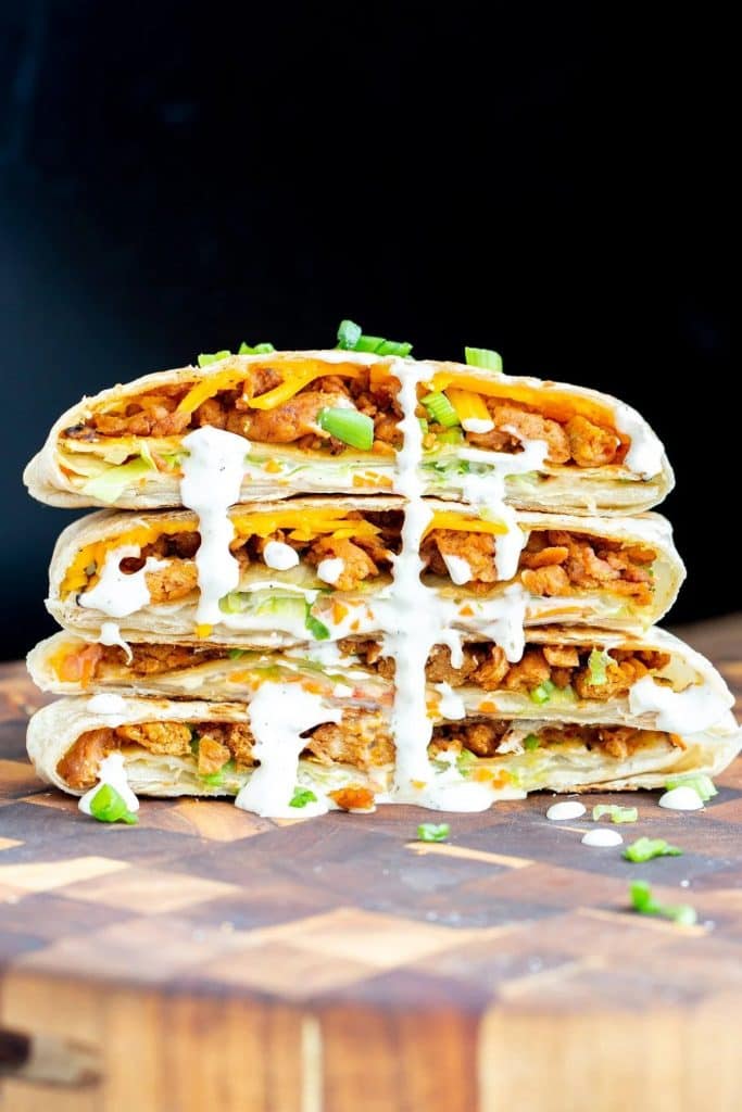 vegan crunch wrap filled with bbq soy curls, carrots, ranch dressing, lettuce, and vegan cheese wrapped in a tortilla. there are four halves stacked on top another, garnished with scallions