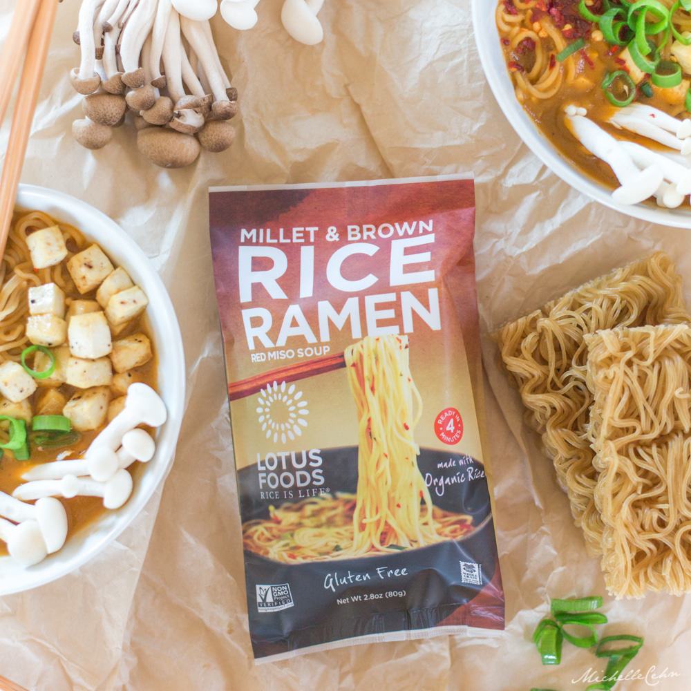 A package of Lotus Foods Millet and Brown Rice Ramen surrounded by bowls of vegan ramen and various ingredients.