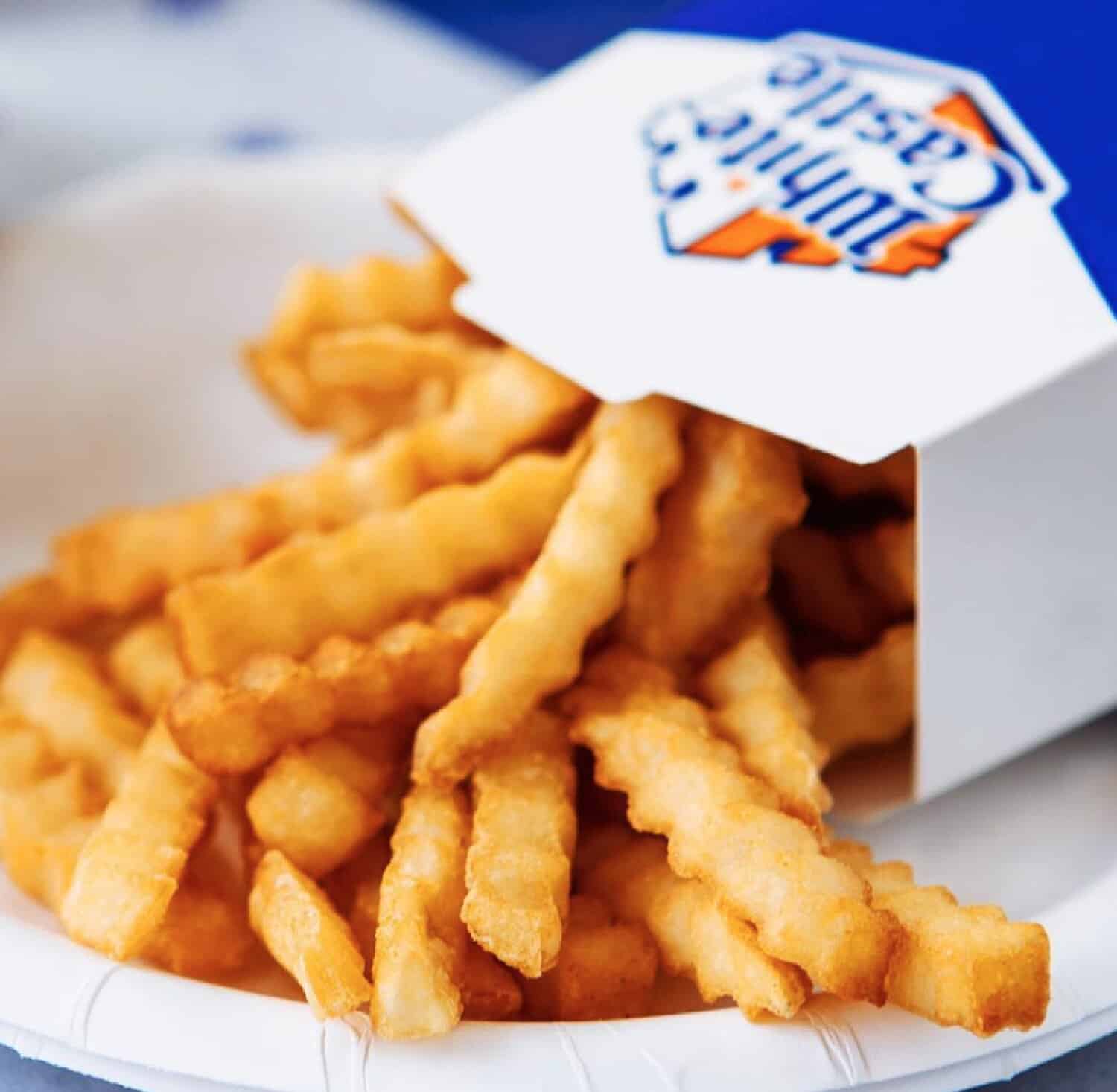 A close up of a box of crinkly fries overturned onto a white plate and spilling out of a blue and white White Castle take out box. 