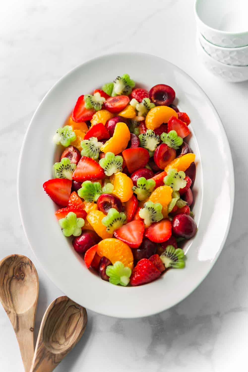 White serving platter with a colorful fruit salad including strawberries, kiwi cut into flower shapes, and mandarin oranges.